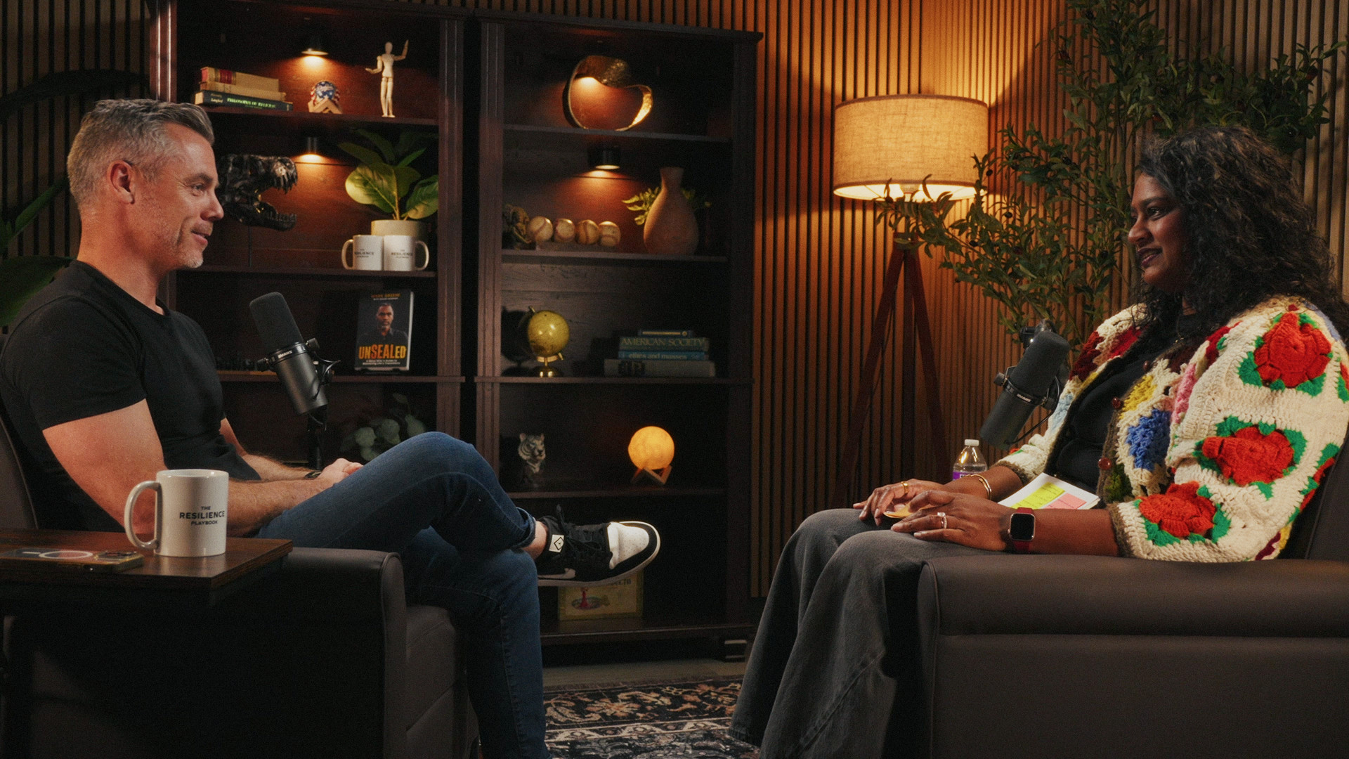 Host and guest talking across the set, bookshelves and a lamp behind them